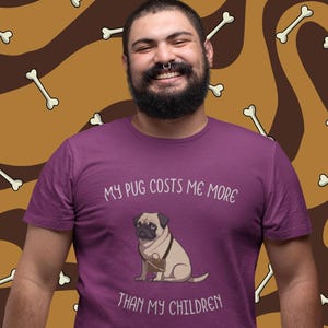 Könnte beinhalten: Ein lila T-Shirt mit dem Text "MY PUG COSTS ME MORE THAN MY CHILDREN" und einer Grafik eines Mops mit einem Korb. Der Hintergrund ist ein Muster aus Knochen auf Braun und Beige.