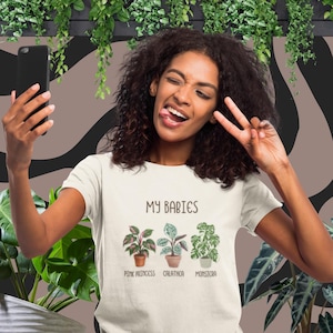 May include: Cream-colored t-shirt with the text "MY BABIES" above illustrations of three potted plants: Pink Princess, Calathea, and Monstera. The person is holding a phone and making a peace sign.