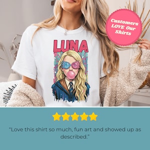 May include: White t-shirt featuring a colorful illustration of a character with the name "LUNA" above. The character is wearing blue and pink glasses and blowing a bubblegum bubble. The shirt has a round neck.