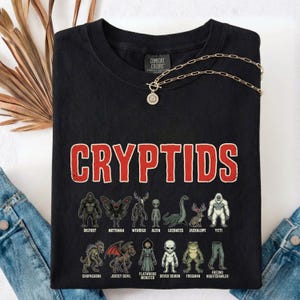 Peut inclure: T-shirt noir avec le mot "CRYPTIDS" en lettres rouges. En dessous, des illustrations de cryptides, dont Bigfoot, Mothman et le monstre du Loch Ness. Un collier en chaîne dorée est posé sur le t-shirt.