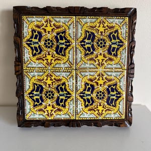 May include: Decorative square tile trivet with four ceramic tiles featuring a yellow, blue, and green floral design. The tiles are set in a dark brown, carved wooden frame. The trivet is a decorative and functional item.