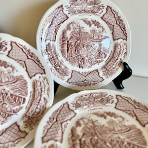May include: Set of three antique white and brown decorative plates. Each plate features a detailed sepia-toned illustration of a ship and figures, with a patterned border and decorative rim. The plates are displayed on a white surface.