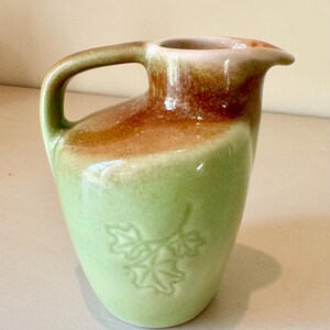 May include: A ceramic pitcher featuring a light green base color that transitions into a warm brown glaze near the top rim. The vessel includes a single handle and a molded relief design of three maple leaves on the front surface of the body.