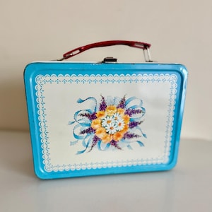 May include: A vintage metal lunchbox with a light blue exterior and a white top panel. The top panel features a floral design with orange, purple, and white flowers, and blue ribbons. The lunchbox has a red handle and a metal clasp.