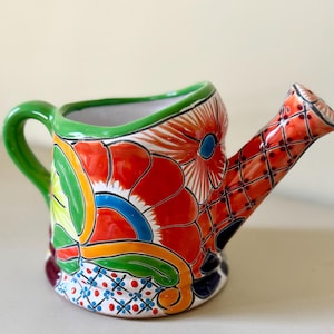 May include: A hand-painted ceramic watering can with a vibrant floral design. The can features a green handle and rim, with a red, orange, and blue floral pattern. The spout has a diamond pattern with red dots. A decorative garden item.