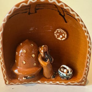 May include: A brown ceramic arch-shaped sculpture featuring a glossy finish with white zigzag trim. Inside, a figure stands beside a large mushroom-shaped structure, holding a small dark animal, with a basket and a circular moon-like accent.