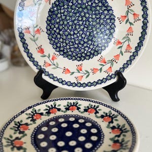 May include: Two ceramic plates with hand-painted patterns. The top plate features a central cluster of small blue and green stars surrounded by a ring of orange bell-shaped flowers. The bottom plate has a dark blue center with white dots.