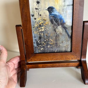 May include: A wooden picture frame with a painting of a blue bird perched on a branch. The frame is made of dark brown wood and has a stand on the back. The painting has a neutral background with yellow and black accents.