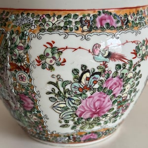 May include: A round ceramic planter featuring a white background with intricate hand-painted details. The design includes vibrant pink flowers, green foliage, colorful butterflies, and a small bird, all framed by an orange and gold decorative border.