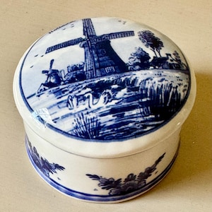 May include: A round, white ceramic trinket box with a blue and white windmill scene on the lid. The box features a detailed landscape with a windmill, swans, and trees. The sides of the box have a blue floral design.