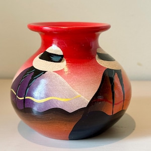 May include: A hand-painted ceramic vase with a vibrant red rim and a rounded body. The vase features a landscape design with mountains in shades of purple, pink, and orange, accented with gold lines. The base is a warm brown.