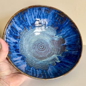 May include: A handmade ceramic bowl with a deep blue glaze. The bowl features a textured surface with shades of blue and brown, and a circular design in the center. The rim has a darker brown color.