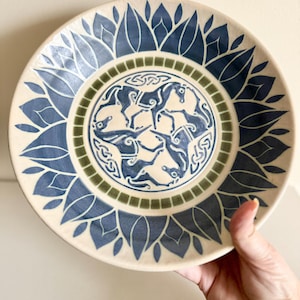 May include: A decorative ceramic plate with a blue and cream color scheme. The plate features a central design of stylized horses within a circular border, surrounded by a leaf-like pattern. The plate is held by a hand.