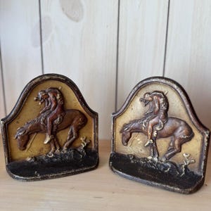 May include: A pair of cast iron bookends featuring a Native American on horseback. The bookends are painted in shades of brown, gold, and black.
