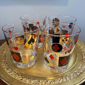 May include: Set of four clear glass tumblers with a gold, black, and red playing card design. The glasses are on a gold tray.