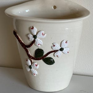 May include: Cream-colored ceramic wall pocket with a curved top and a hole for hanging. The front features a raised relief design of white dogwood flowers with red accents and green leaves on a brown branch.