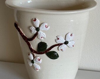 May include: Cream-colored ceramic wall pocket with a curved top and a hole for hanging. The front features a raised relief design of white dogwood flowers with red accents and green leaves on a brown branch.