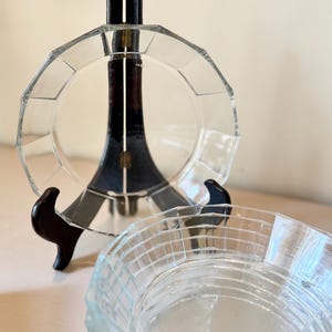 May include: A clear glass plate with a faceted, geometric rim displayed on a dark wooden stand, positioned behind a matching clear glass bowl that features a textured, grid-like pattern on its exterior surface and a smooth, circular base.