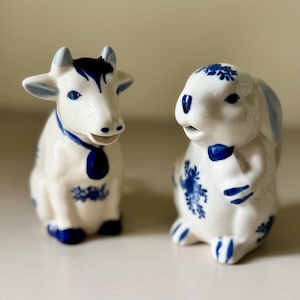 May include: Two ceramic figurines: a cow and a rabbit. The cow is white with blue accents, including horns, a collar, and floral patterns. The rabbit is also white with blue details, featuring a bow tie and floral designs. Both have a glossy finish.