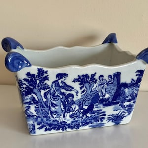 May include: A rectangular, white ceramic planter with a blue and white pastoral scene. The planter has a scalloped rim and blue, teardrop-shaped handles. The blue design depicts figures, trees, and a bridge.