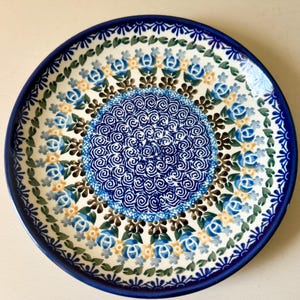 May include: A round ceramic plate with a dark blue rim and a floral pattern. The plate features a central blue spiral design surrounded by blue and yellow flowers and green leaves on a white background.