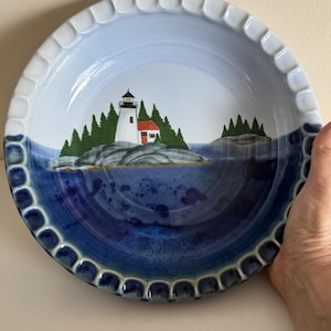 May include: A ceramic plate with a lighthouse scene. The plate features a white lighthouse with a red roof on a rocky island, surrounded by green trees and blue water. The rim has a scalloped edge.