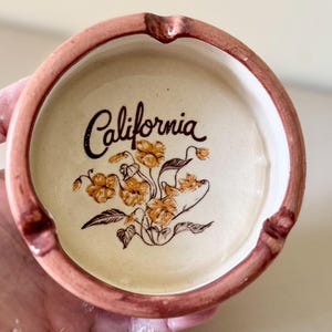 May include: A round, ceramic ashtray with a light beige interior and a brown rim. The word "California" is written in cursive script, with a floral design in shades of orange and brown.