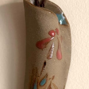 May include: A handmade ceramic wall hanging with a brown cord. The ceramic piece is a curved shape with a beige base color. It features pink and blue floral designs. The piece is likely for home decor.