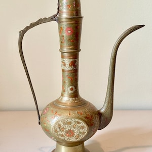 May include: A tall brass pitcher with a long, curved spout and a handle. The surface features intricate etched floral patterns with inlaid red, green, and white enamel details. It has a bulbous body, a slender neck, and a flared base.