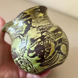 May include: A small, handmade ceramic vase with a unique design. The vase features a dark brown base color with a striking pattern of lime green and yellow swirls. The vase has a rounded body and a slightly flared rim.