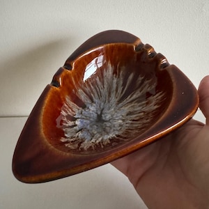 May include: A triangular ashtray with a glossy, brown exterior and a central design of white, gray, and blue brushstrokes. The ashtray has a smooth, curved rim and a small notch on each corner.