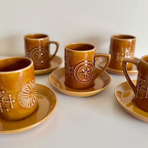 May include: Set of six brown ceramic mugs with matching saucers. Each mug features a handle and embossed geometric designs, including starburst and cross-like patterns. The mugs and saucers have a glossy finish.