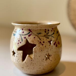 May include: A speckled beige ceramic candle holder with star cutouts. The candle holder has a round shape with a slightly flared rim. It is decorated with painted green leaves and red berries.