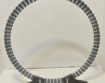 Può includere: Oggetto decorativo circolare grigio e blu con un motivo ripetuto. L'oggetto presenta una serie di blocchi rettangolari grigi con accenti blu e bianchi. Due supporti neri angolari sono in basso, ciascuno con un simbolo bianco.