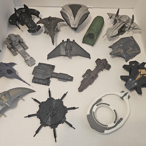 May include: A collection of various toy spacecraft models in shades of gray, black, and silver. The models feature intricate designs, including wings, fins, and detailed surface textures. Some models have a metallic finish, while others are matte. The toys are arranged on a white surface.