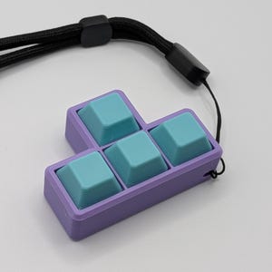 May include: A purple and teal keycap set in a Tetris block shape, attached to a black lanyard. The keycaps are teal, and the frame is purple. The lanyard is black and has a black plastic clasp.