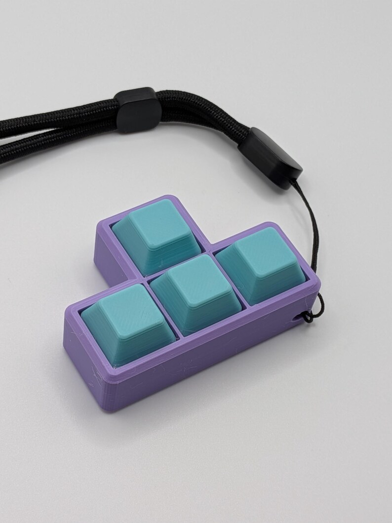 May include: A purple and teal keycap set in a Tetris block shape, attached to a black lanyard. The keycaps are teal, and the frame is purple. The lanyard is black and has a black plastic clasp.