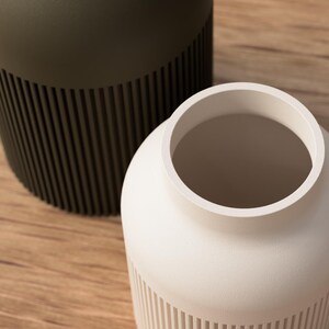 May include: Two modern vases, one in a matte olive green and the other in a creamy white. Both have a cylindrical shape with vertical ribbed detailing on the lower portion. The vases sit on a light brown wooden surface.
