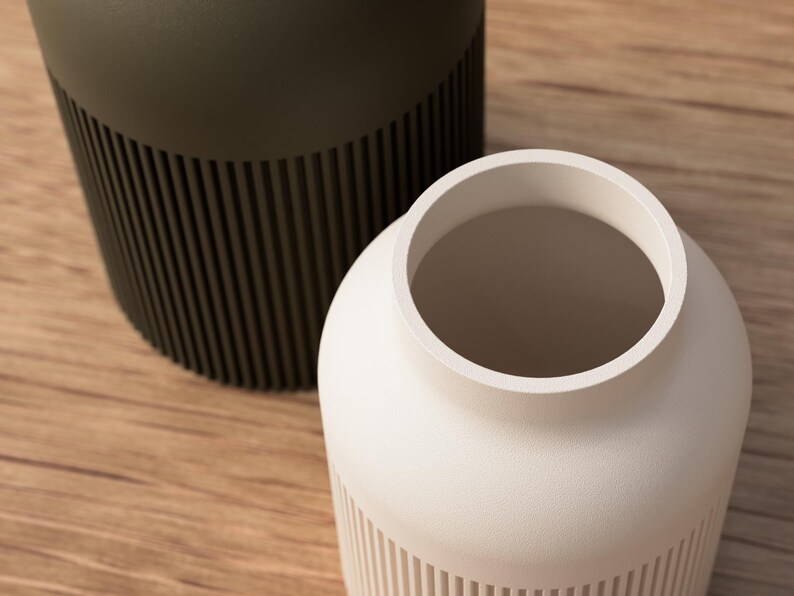 May include: Two modern vases, one in a matte olive green and the other in a creamy white. Both have a cylindrical shape with vertical ribbed detailing on the lower portion. The vases sit on a light brown wooden surface.