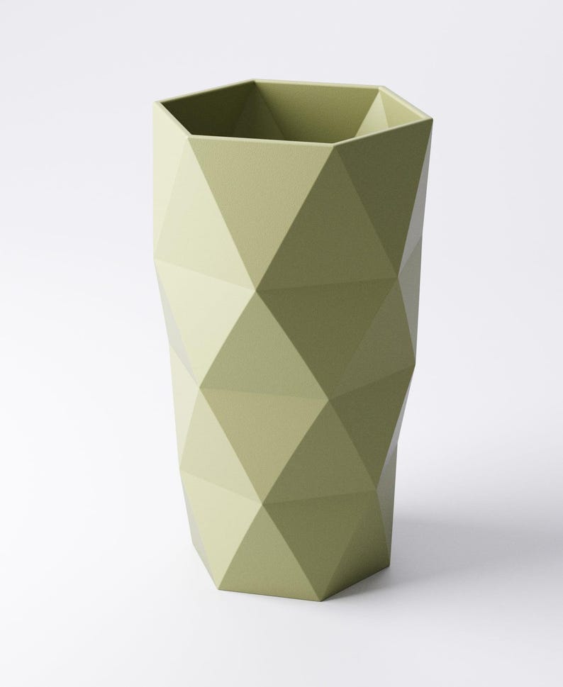 Op de afbeelding: Een saliegroene, geometrische vaas met een facetontwerp. De vaas heeft een zeshoekige bovenkant en een reeks ruitvormige facetten die een textuur cre&euml;ren. De vaas is effen van kleur en staat rechtop tegen een witte achtergrond.