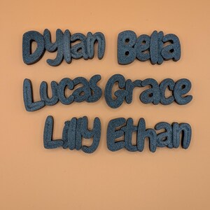 May include: Six dark grey, 3D-printed name cutouts: Dylan, Bella, Lucas, Grace, Lilly, and Ethan. The names are in a playful, rounded font, set against an orange background. Suitable for personalised decor or craft projects.