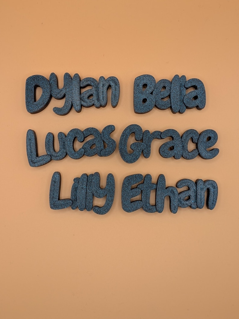 May include: Six dark grey, 3D-printed name cutouts: Dylan, Bella, Lucas, Grace, Lilly, and Ethan. The names are in a playful, rounded font, set against an orange background. Suitable for personalised decor or craft projects.