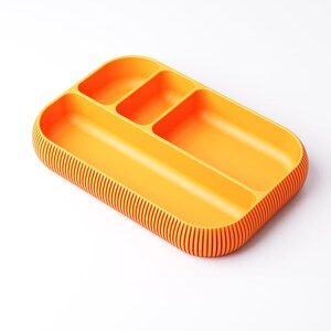 May include: An orange rectangular tray with three compartments and a ribbed edge. The tray is made of a solid, opaque material and is designed for organizing items. The compartments are different sizes.
