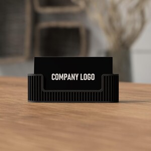May include: A black business card holder with a vertical ribbed design. The holder displays a black business card with the text "COMPANY LOGO" in white. The holder sits on a wooden surface.