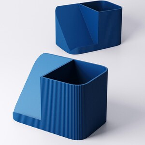 May include: Two blue desk organizers. Each organizer has a rectangular shape with a sectioned interior. One section is a tall, open container, and the other is a lower, angled compartment. The organizers have a ribbed texture on the sides.