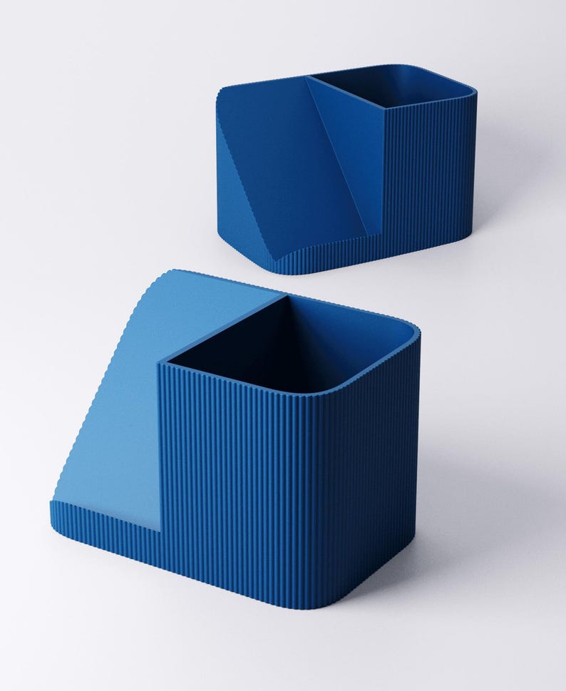 May include: Two blue desk organizers. Each organizer has a rectangular shape with a sectioned interior. One section is a tall, open container, and the other is a lower, angled compartment. The organizers have a ribbed texture on the sides.