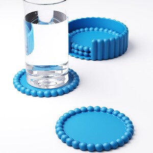 May include: A clear glass of water sits on a round, light blue coaster with a beaded edge. Two additional coasters are visible, one with a similar design and another with a vertical ribbed design. The coasters are made of a flexible material.
