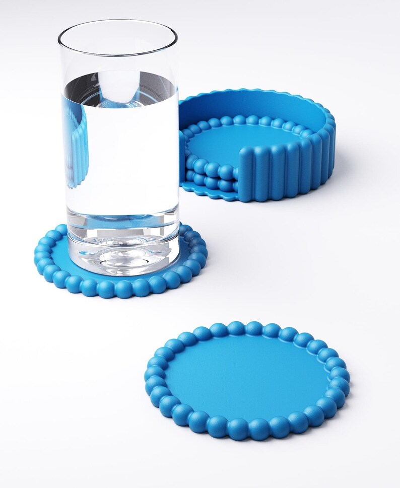 May include: A clear glass of water sits on a round, light blue coaster with a beaded edge. Two additional coasters are visible, one with a similar design and another with a vertical ribbed design. The coasters are made of a flexible material.