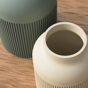 May include: Two ceramic vases. One is a light sage green color, and the other is off-white. Both vases have a cylindrical shape with vertical ribbed detailing on the lower half. The vases are on a wooden surface.