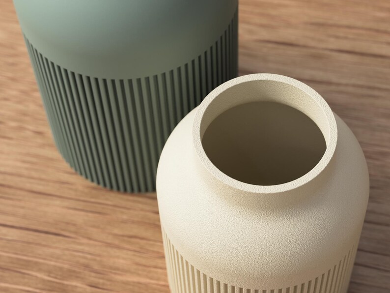 May include: Two ceramic vases. One is a light sage green color, and the other is off-white. Both vases have a cylindrical shape with vertical ribbed detailing on the lower half. The vases are on a wooden surface.